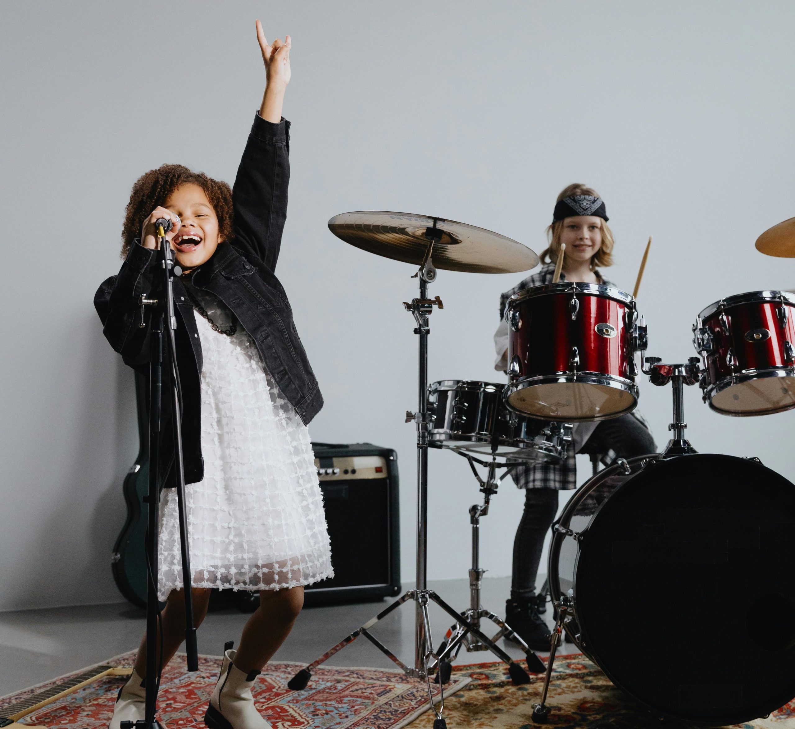 two kids in a rockband. young girl has her hand in the air pointing finger singing into a microphone whole young boy dressed like a rockstar plays the drum kit
